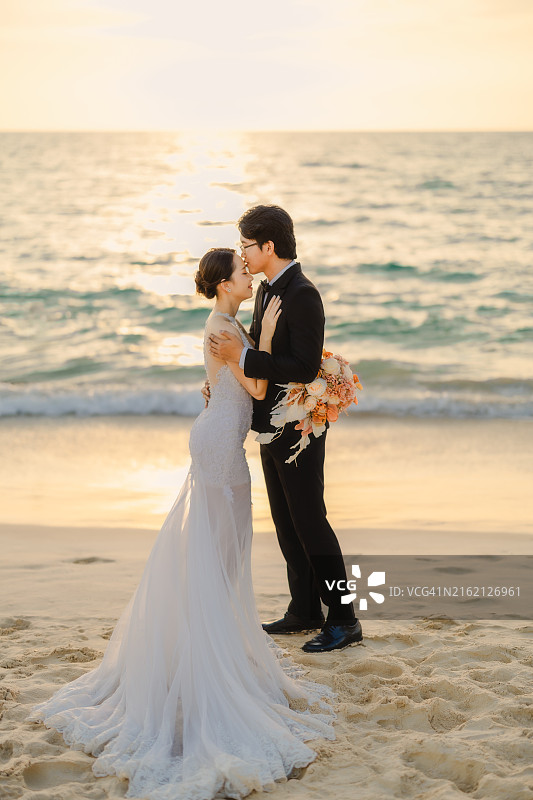 新郎身穿西装，新娘身穿白色连衣裙，手捧鲜花，面带微笑地站在日落时分的海滩上图片素材