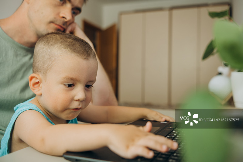 坐在父亲腿上，尝试在家工作的小男孩图片素材