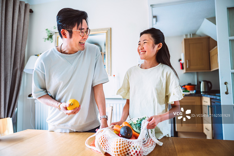 年轻的亚洲父亲和十几岁的女儿在家一起从食品袋中取出新鲜水果和蔬菜图片素材