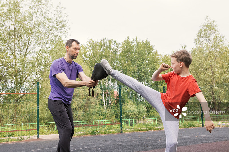 户外运动场上教练和男孩之间的武术训练-使用脚垫进行高踢腿练习图片素材
