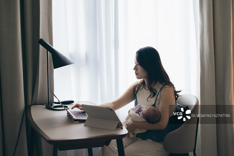 亚洲母亲在笔记本电脑上处理工作任务和家庭财务，同时照顾她的宝宝，体现了兼顾工作和育儿的概念图片素材