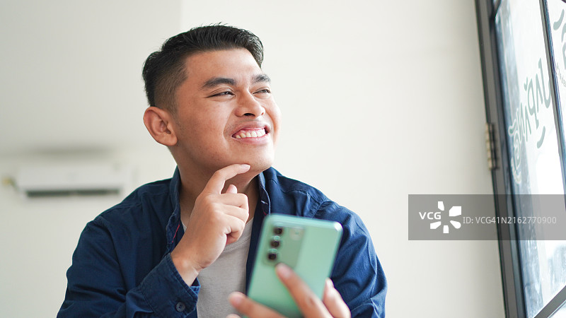 使用智能手机思考并微笑的亚洲男人图片素材