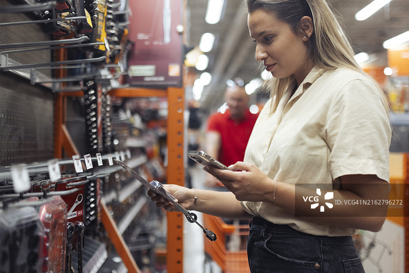 在商店里购买工具的无忧无虑的女人侧面，她正在看手机上的工具图片，以确保不会买错图片素材