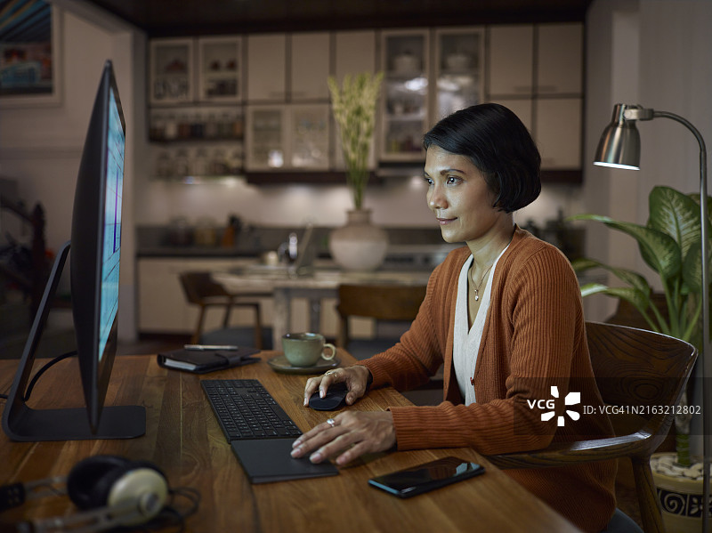 中年亚洲女人在家里的办公室工作，晚上使用台式电脑，背景是厨房图片素材