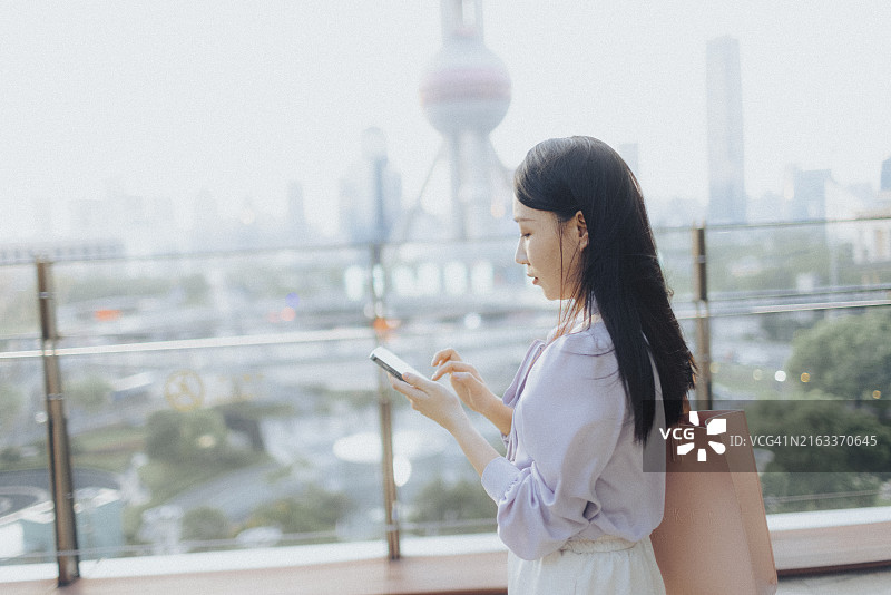 在市中心陆家嘴金融区金融摩天大楼前户外用智能手机发短信的年轻漂亮亚洲女性图片素材