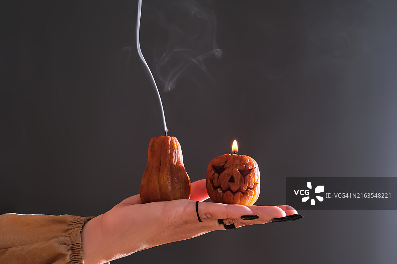做黑色美甲、戴戒指的女人手拿两支点燃的万圣节南瓜造型蜡烛。照片展示了蜡烛熄灭后的大气白色烟雾。图片素材