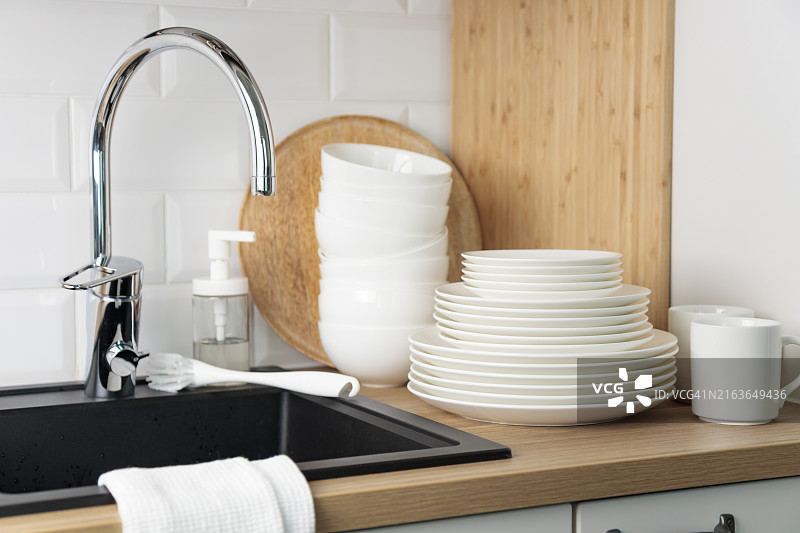 一套干净的白色盘子、餐具，靠近黑色厨房水槽，水槽配有木制柜台上镀铬水龙头和白色地铁瓷砖挡板，配有洗碗液、刷子和海绵图片素材