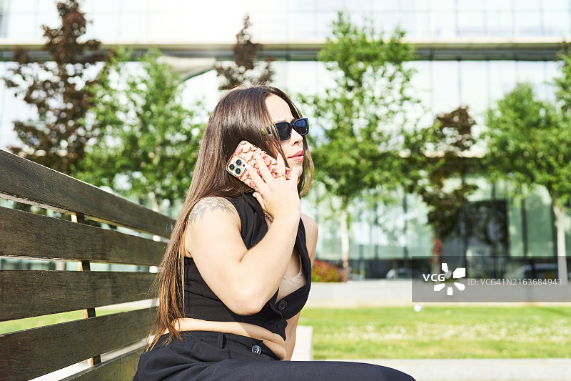 穿着讲究的漂亮女人坐在建筑物外的长椅上打电话，侧视图图片素材