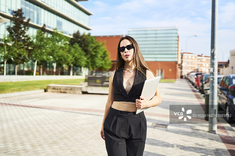 戴着太阳镜，在建筑物外拿着笔记本电脑看着镜头的女人肖像，正面图图片素材
