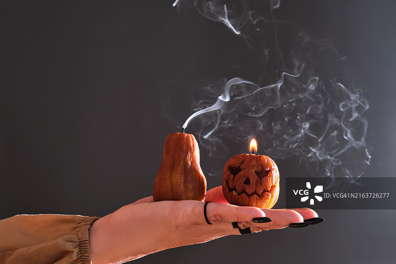 做黑色美甲、戴戒指的女人手拿两支点燃的万圣节南瓜造型蜡烛。照片展示了蜡烛熄灭后的大气白色烟雾。图片素材
