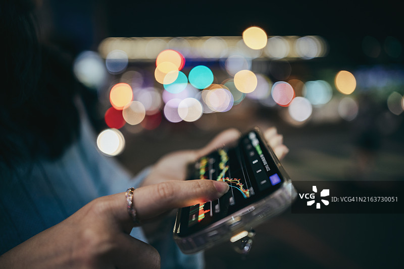 夜晚城市街道，女性手持手机浏览股市图表，背景虚化图片素材