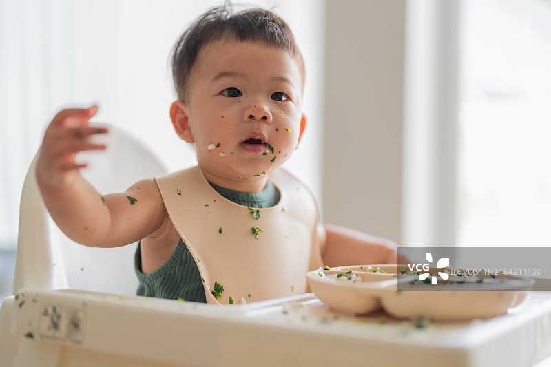 可爱的亚洲男婴独自坐在高脚椅上吃饭，母亲在家用勺子喂他，婴儿成长里程碑图片素材