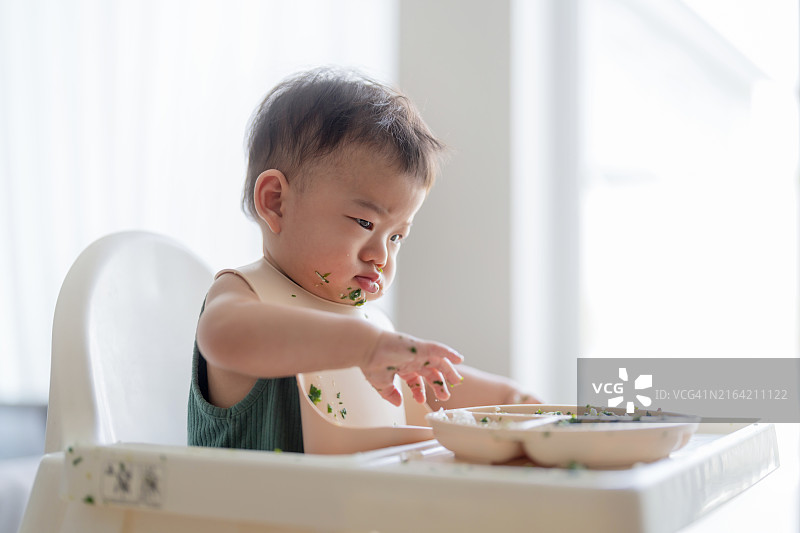 可爱的亚洲男婴独自坐在高脚椅上吃饭，母亲在家用勺子喂他，婴儿成长里程碑图片素材