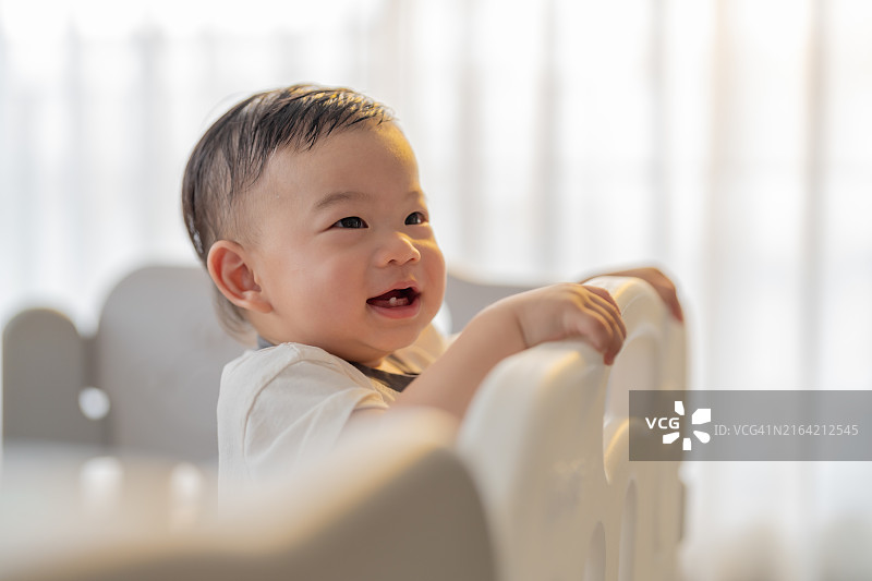 可爱的亚洲男婴（8-12个月）在游戏围栏厨房里享受站立，拿着白色围栏，在家里的游戏围栏里玩玩具，室内背景，阳光明媚，有趣的笑声，这个男孩真可爱图片素材