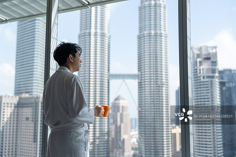 男子穿着浴袍喝着咖啡，欣赏着早晨摩天大楼的景色图片素材