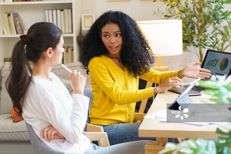 不同国籍的两位初创女商人身穿休闲装，在家互相帮助，使用笔记本电脑、平板电脑和纸张开启小生意图片素材