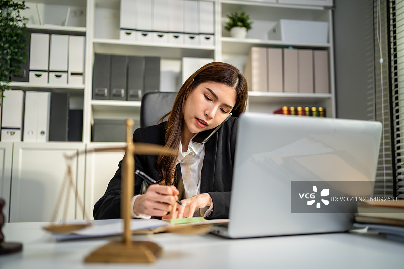 亚洲女律师在法庭上用手机交谈和工作。有魅力的法官律师在办公室的桌旁查看纸质文件。法律和法律商业服务概念图片素材