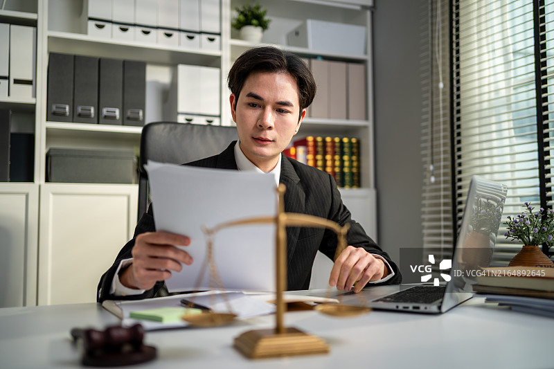 亚洲男律师在法庭上使用锤子和正义天平工作，有魅力的法官律师坐在桌旁，看着办公室里的纸质文件，法律和法律商业服务概念图片素材