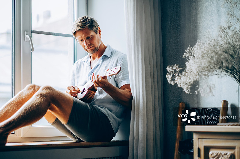 男人坐在窗边弹尤克里里，享受室内宁静时刻图片素材
