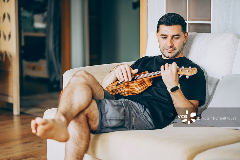 年轻男子舒适地坐在沙发上弹奏尤克里里，享受家中宁静的时刻图片素材