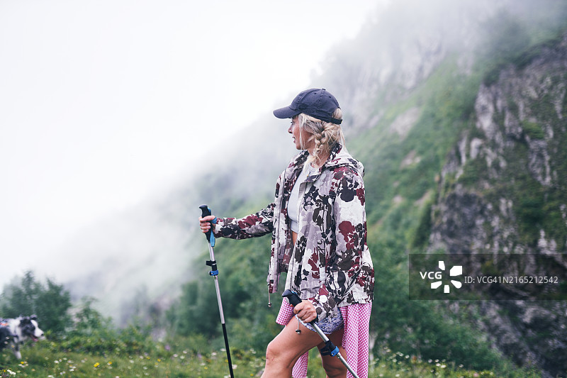 女子在雾气缭绕的山坡上徒步旅行，身穿迷彩夹克和粉色裙子，周围是茂密的植被和雾蒙蒙的气氛。图片素材