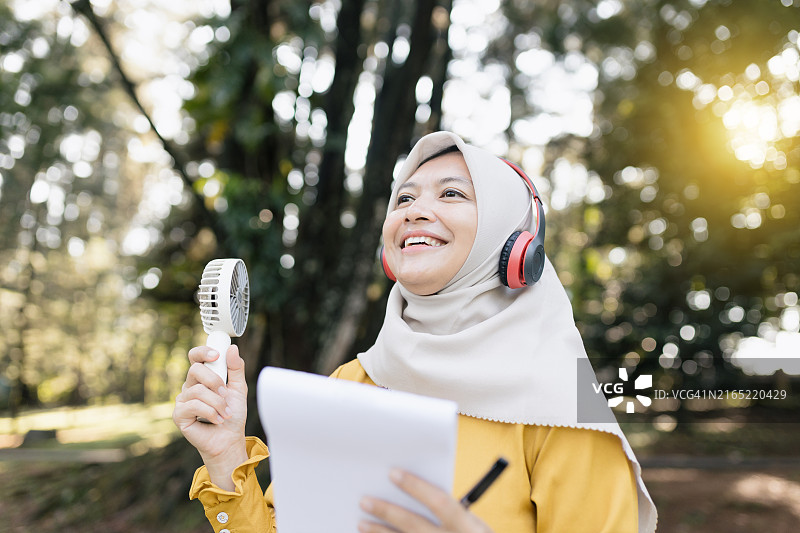 穆斯林妇女戴着无线耳机，拿着笔记本在公园野餐时工作图片素材