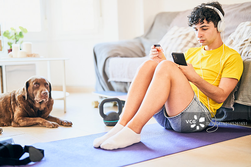 年轻男人坐在瑜伽垫上听音乐，并使用智能手机在网上购物图片素材