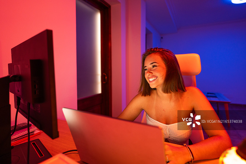 在智能彩色灯光下晚上用笔记本电脑微笑工作的年轻女人图片素材