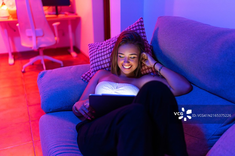 年轻女人在家使用平板电脑在沙发上放松，有智能灯图片素材