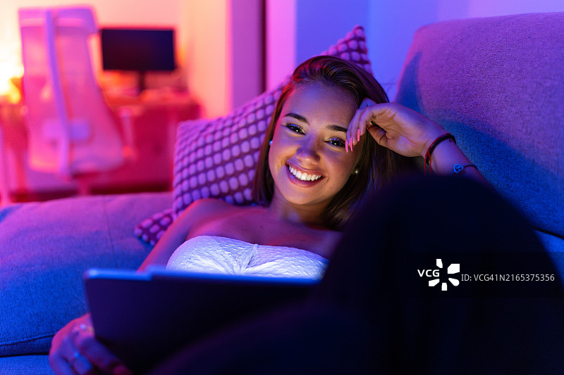 年轻女人在沙发上使用平板电脑，彩色灯光下放松身心图片素材