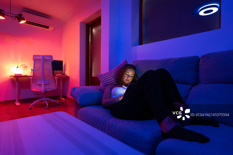 年轻女人在家中彩色灯光下在沙发上用平板电脑放松图片素材