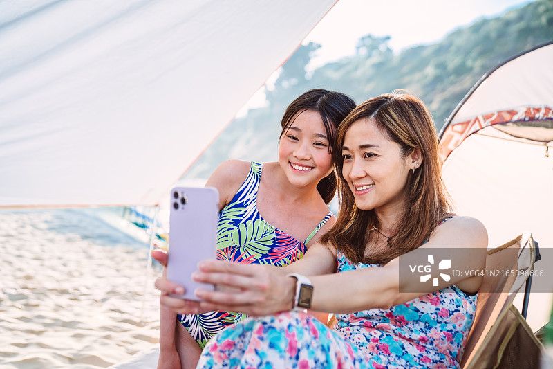 年轻的亚洲母亲和十几岁的女儿在阳光明媚的海滩上用智能手机自拍图片素材