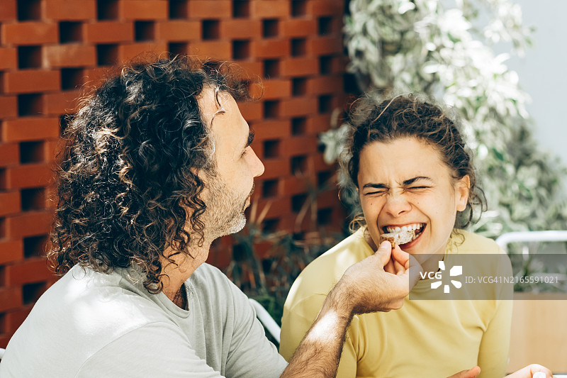 快乐的男人在户外咖啡馆享受阳光明媚的一天，喂迷人的女人一片面包，逗她发笑。家人共度美好时光。图片素材