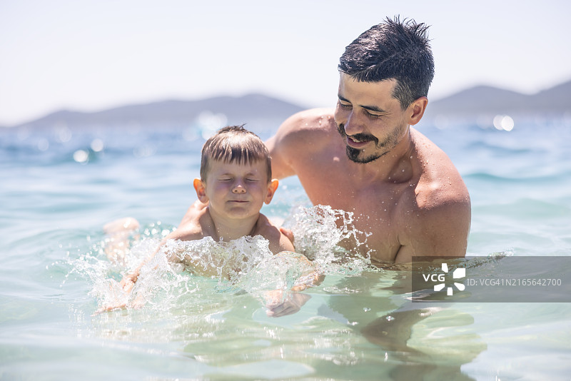 快乐的孩子和他的父亲在日落时在海边或湖边游泳，玩得很开心。爸爸教他小儿子游泳。在度假胜地夏季，家庭与儿童的积极假期图片素材