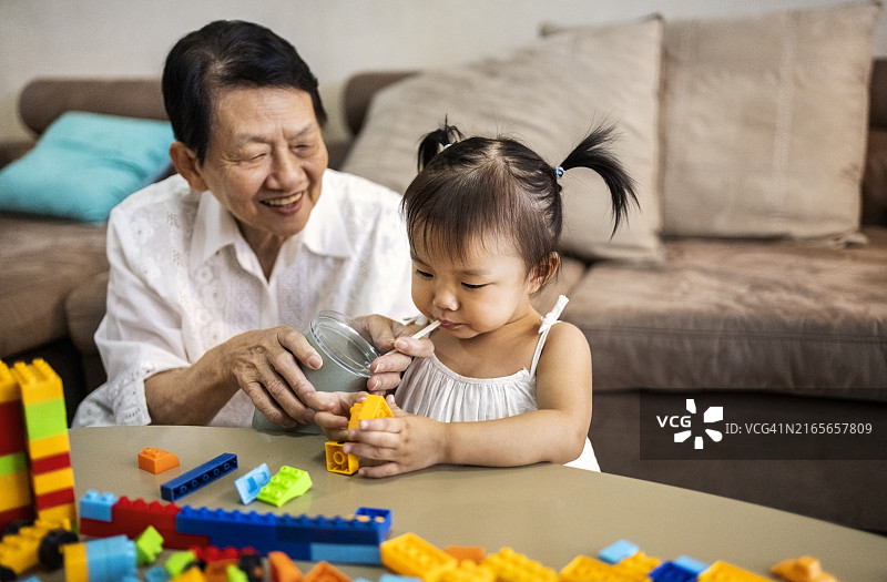 在客厅里和奶奶一起玩积木喝牛奶的可爱小女孩图片素材