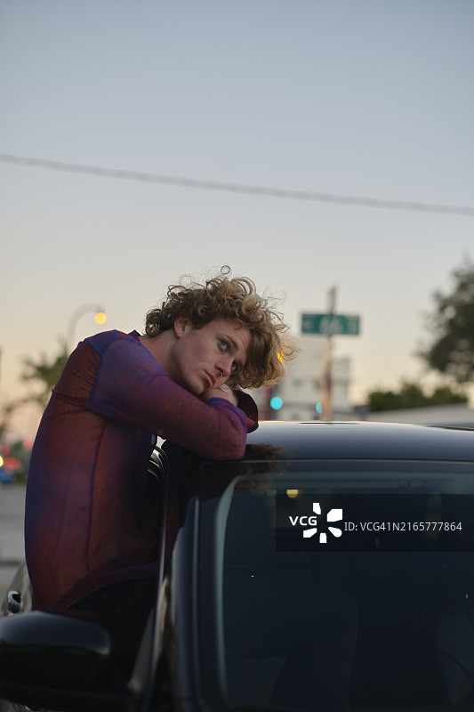 黄昏时分，倚靠在汽车上的卷发年轻男子，背景是城市灯光图片素材