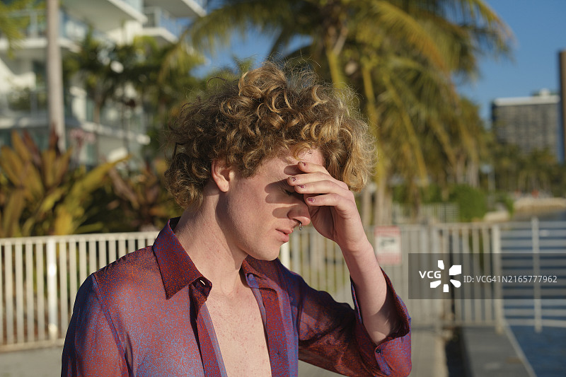 在阳光下眯着眼睛的卷发男人，背景是海滨棕榈树和建筑物图片素材