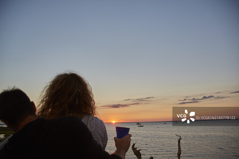 一对夫妇在海边欣赏日落，女人拿着饮料，男人搂着她的肩膀图片素材