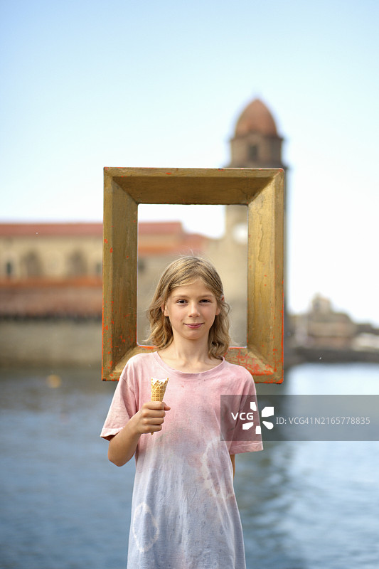 微笑的儿童拿着冰淇淋站在空相框后，背景是历史建筑和水面图片素材