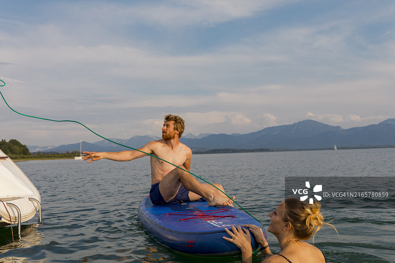 男人坐在滑水板上，平静的湖面上，女人从船上递给他拖绳，背景是山图片素材