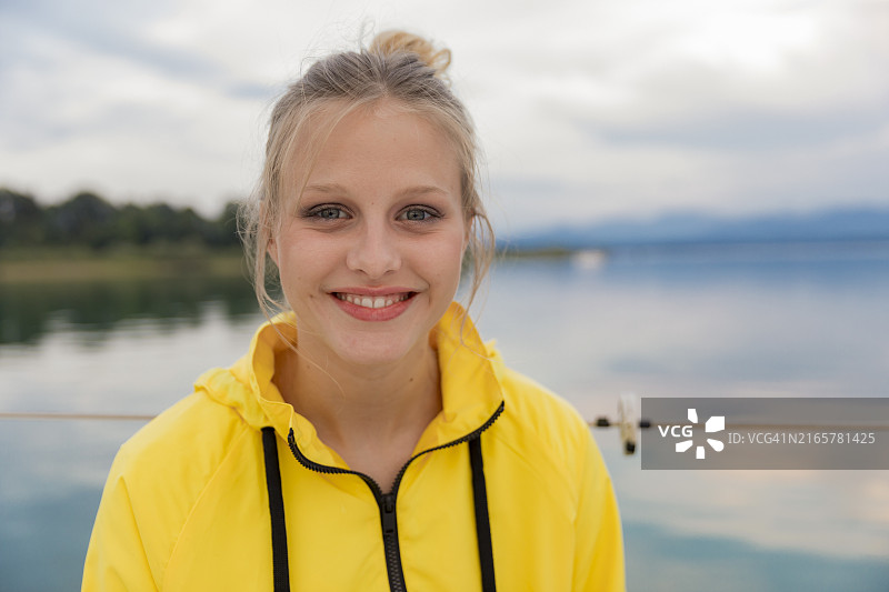 微笑的年轻女人，身穿黄色夹克，站在平静的湖边，背景是多云的天空图片素材