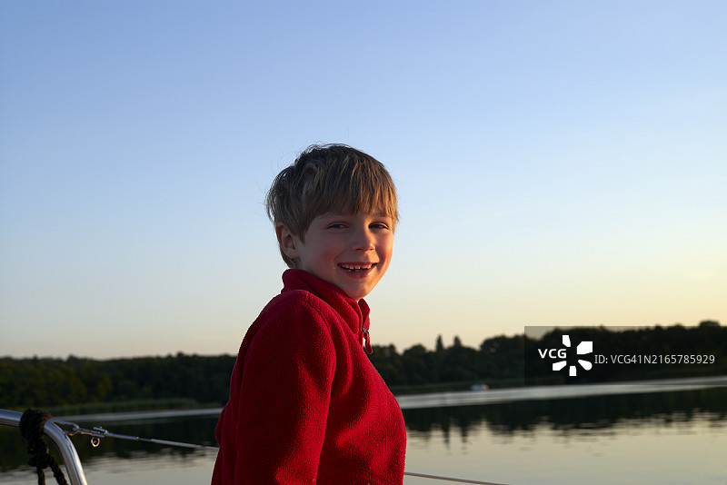 微笑的男孩穿着红色连帽衫欣赏湖边日落图片素材