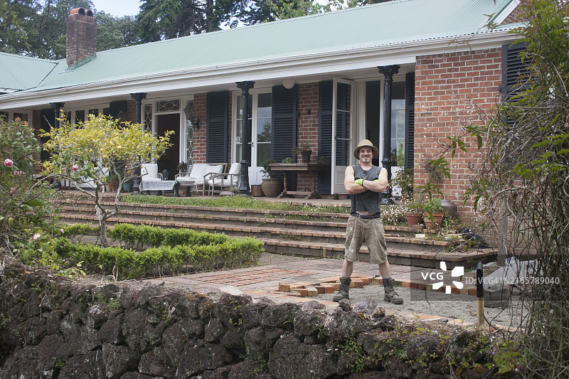 一位快乐的房主在新西兰奥克兰，站在拥有迷人门廊和精心维护的花园的美丽房子前，双手交叉图片素材