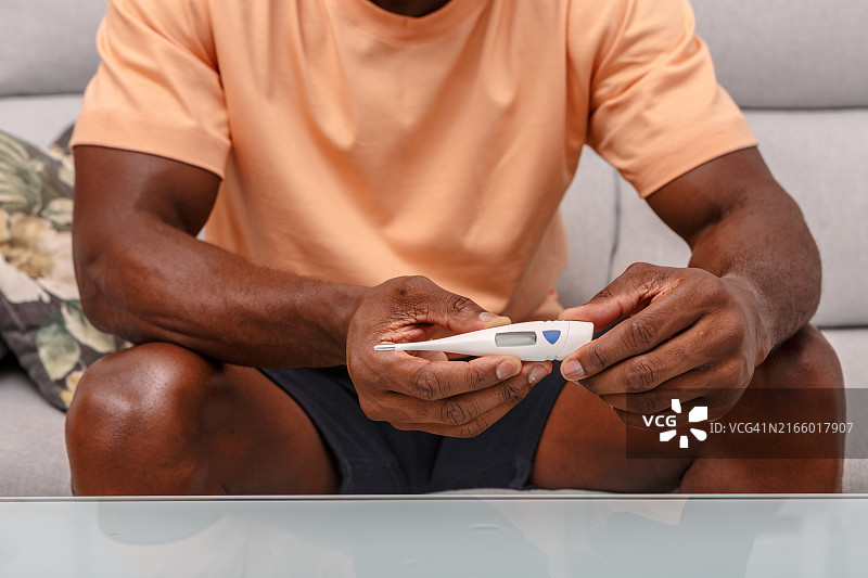 生病在家坐在沙发上用电子温度计测量体温的男人图片素材