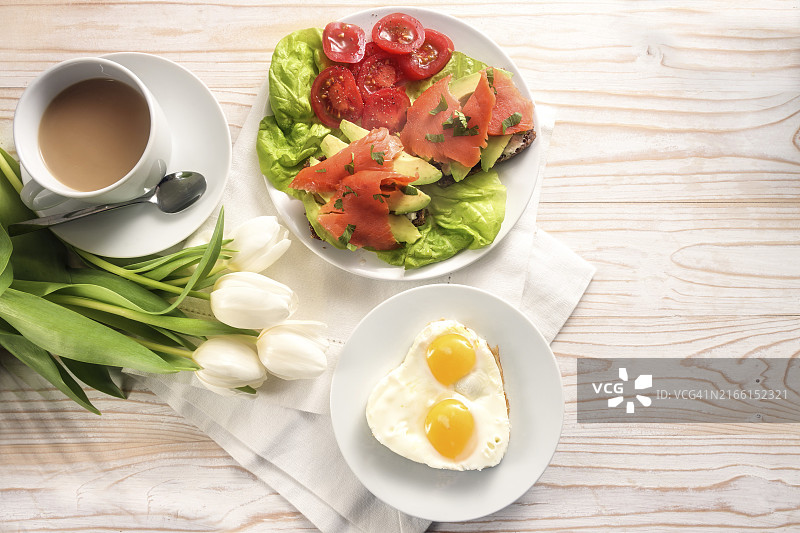 充满爱意的健康早餐：心形煎蛋、牛油果烟熏三文鱼三明治配生菜番茄沙拉、咖啡和白色郁金香，明亮木桌背景。图片素材