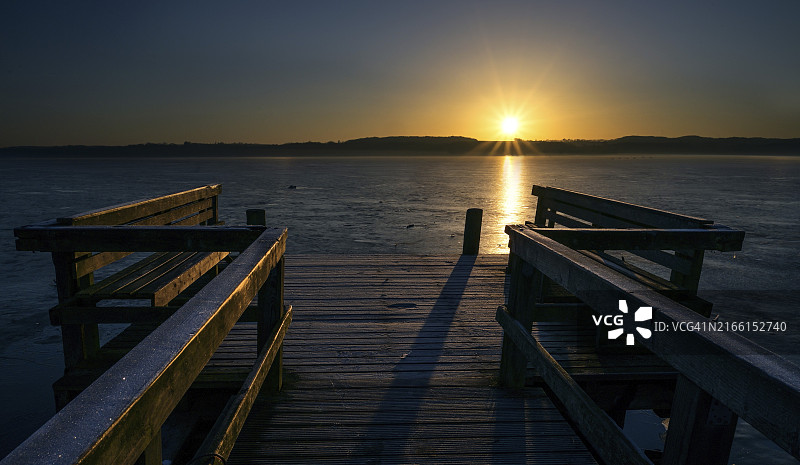 金色朝阳照耀下，冰冻湖面上带有长椅的木制码头图片素材
