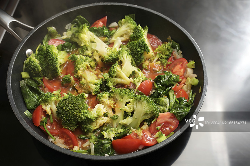炉子上放着西兰花、西红柿、葱和菠菜的蔬菜平底锅，用新鲜食材烹饪健康的素食，选定焦点，窄景深图片素材