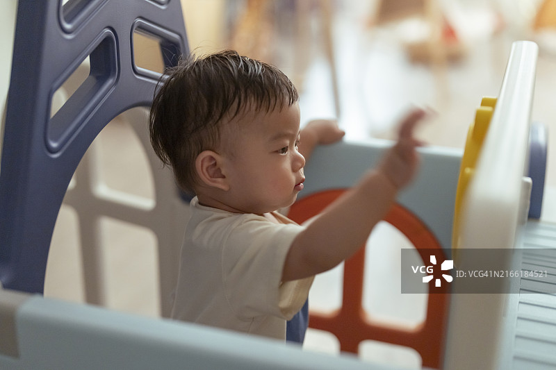 可爱的亚洲男婴（8-12个月）在游戏围栏厨房里享受站立，拿着白色围栏，在家里的游戏围栏里玩玩具，室内背景，阳光明媚，有趣的笑声，这个男孩真可爱图片素材
