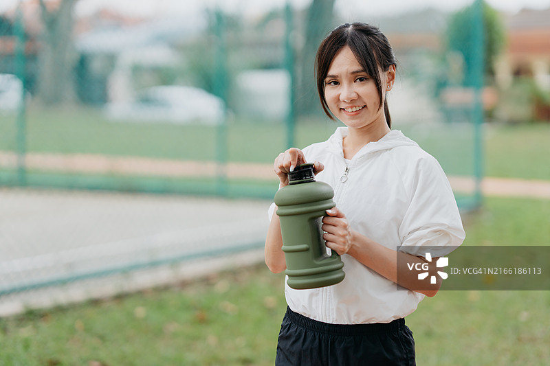 亚洲女性运动员在城市公园的场地上喝着瓶装新鲜水，休息一下锻炼后的疲劳。图片素材