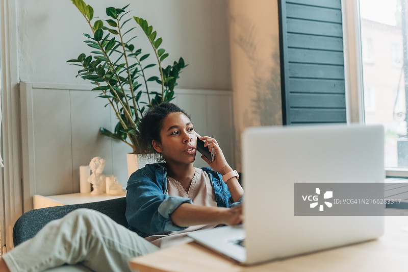 快乐的非洲裔女学生坐在桌旁，使用笔记本电脑读书学习，课间休息时拿着手机上网，收到朋友发来的消息，聊着周末的计划图片素材